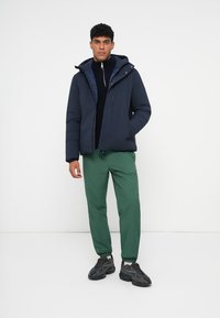 Marineblå jakke med hætte, lynlås foran og elastiske manchetter, parret med grønne tapered bukser og sorte sneakers på hvid baggrund.