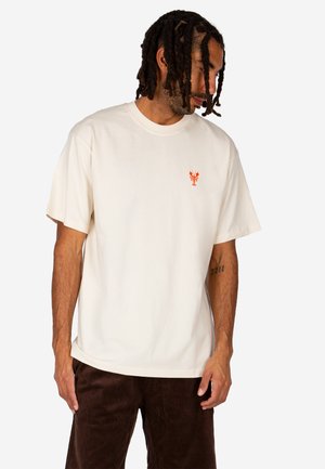 Mann mit Dreadlocks trägt ein cremefarbenes T-Shirt mit kurzen Ärmeln und kleinem orangefarbenem Hummer-Logo, blickt nach unten, kombiniert mit dunkelbrauner Hose.