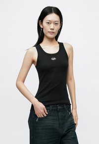 Fekete ujjatlan trikó kerek nyakkivágással és egy kis logóval a közepén. Az anyag sima és testhezálló, sötét farmernadrággal párosítva.