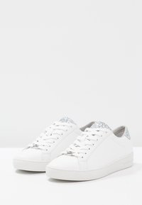 Sneakers bianchi a bassa punta realizzati in pelle, con un accento argentato scintillante sul tallone, punta rotonda e suola piatta in gomma.