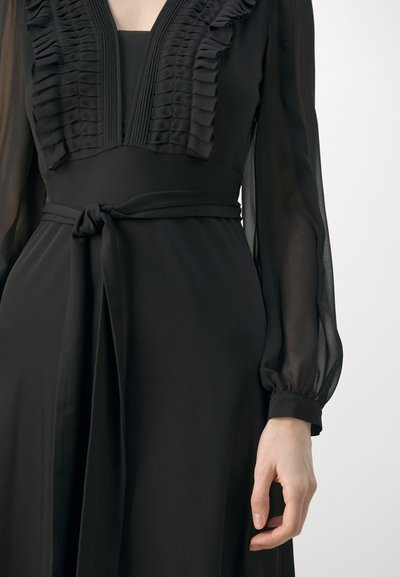 Robe noire avec un détail de décolleté à volants, des manches longues en voile et une taille à nouer. Tissu lisse avec un design fluide.
