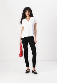 Bílá košile s výstřihem do V, černé slim jeans, černé boty a červená křížová taška. Model stojí před jednoduchým bílým pozadím.