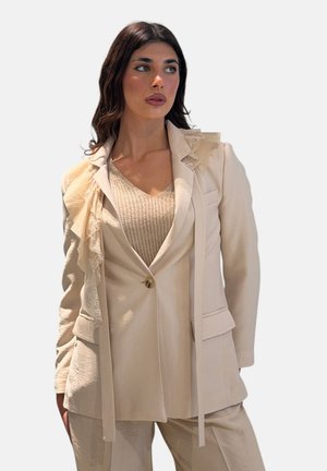 Donna che indossa un blazer beige su misura con dettagli in pizzo e volant sopra un top lavorato a maglia e pantaloni coordinati, guarda di lato su sfondo semplice.