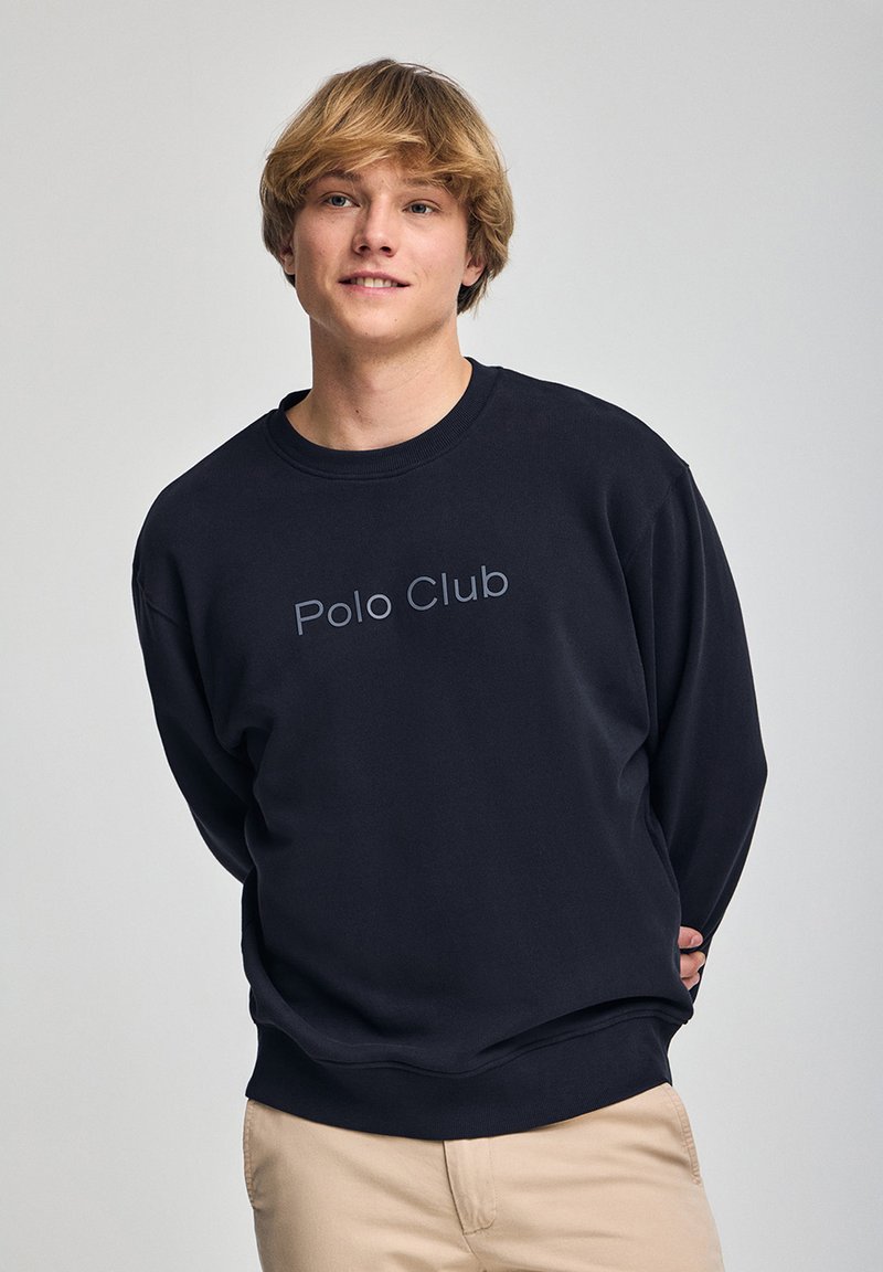 Sudadera azul marino con cuello redondo y puños acanalados. Presenta un logo de texto "Polo Club" en gris a lo largo del pecho. Textura suave, parecida al forro polar.
