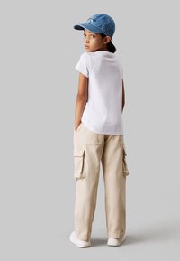 Camiseta blanca de cuello redondo, pantalones cargo beige con bolsillos laterales y gorra de mezclilla azul. Zapatillas blancas completan el conjunto. Vista trasera.