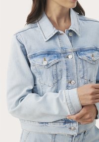 Svetlo modrá denimová bunda s dvoma vreckami na hrudi, striebornými gombíkmi a prešitou aplikáciou. Tkanina je mäkká s klasickým strihom a golierom.