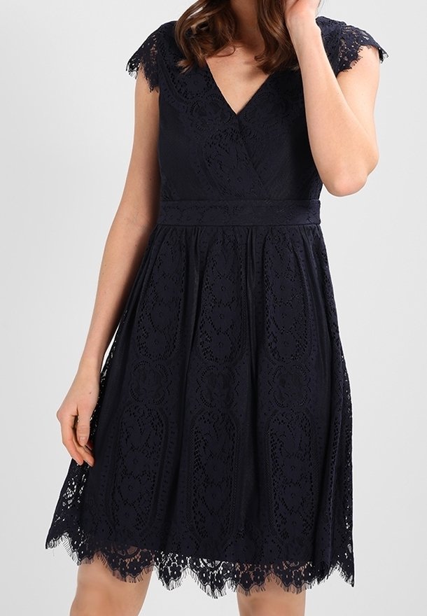 Femme portant une robe noire en dentelle longueur genou avec des manches courtes et un décolleté en V, debout devant un fond clair uni.