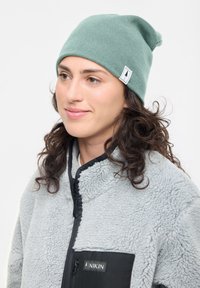 Green beanie hat made of stretchy fabric with a slouchy design. Features a small logo tag. Worn with a grey, fluffy fleece jacket.