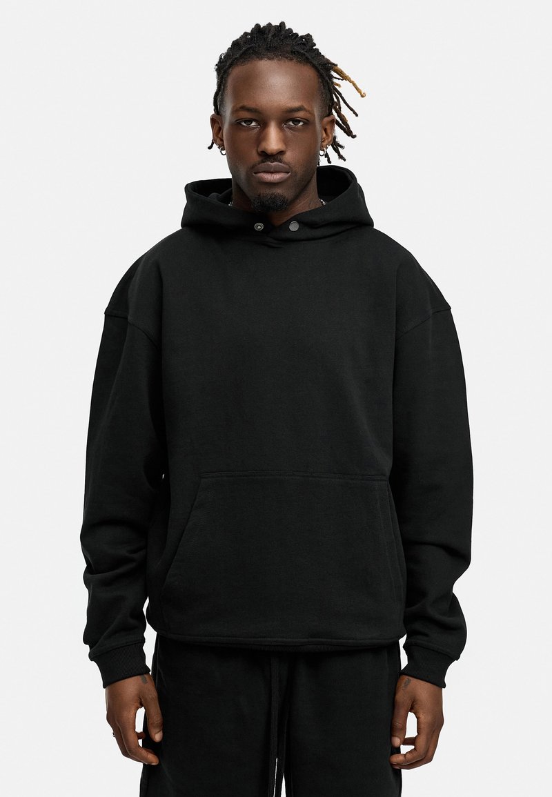 Black oversized hoodie in soft fabric, featuring a kangaroo pocket, drawstring hood, and snap closures at the neck. Smooth texture, relaxed fit.