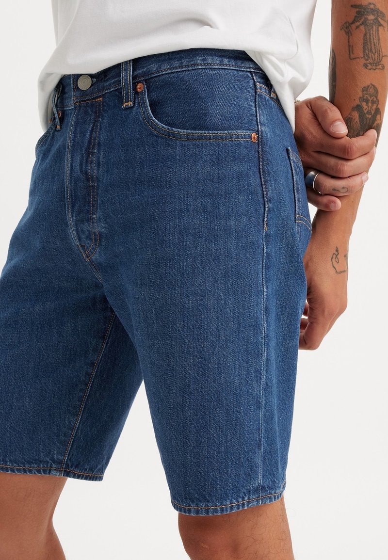 Homme portant un short en jean bleu et une chemise blanche tenant un bras tatoué avec une bague en argent à son doigt sur fond blanc.