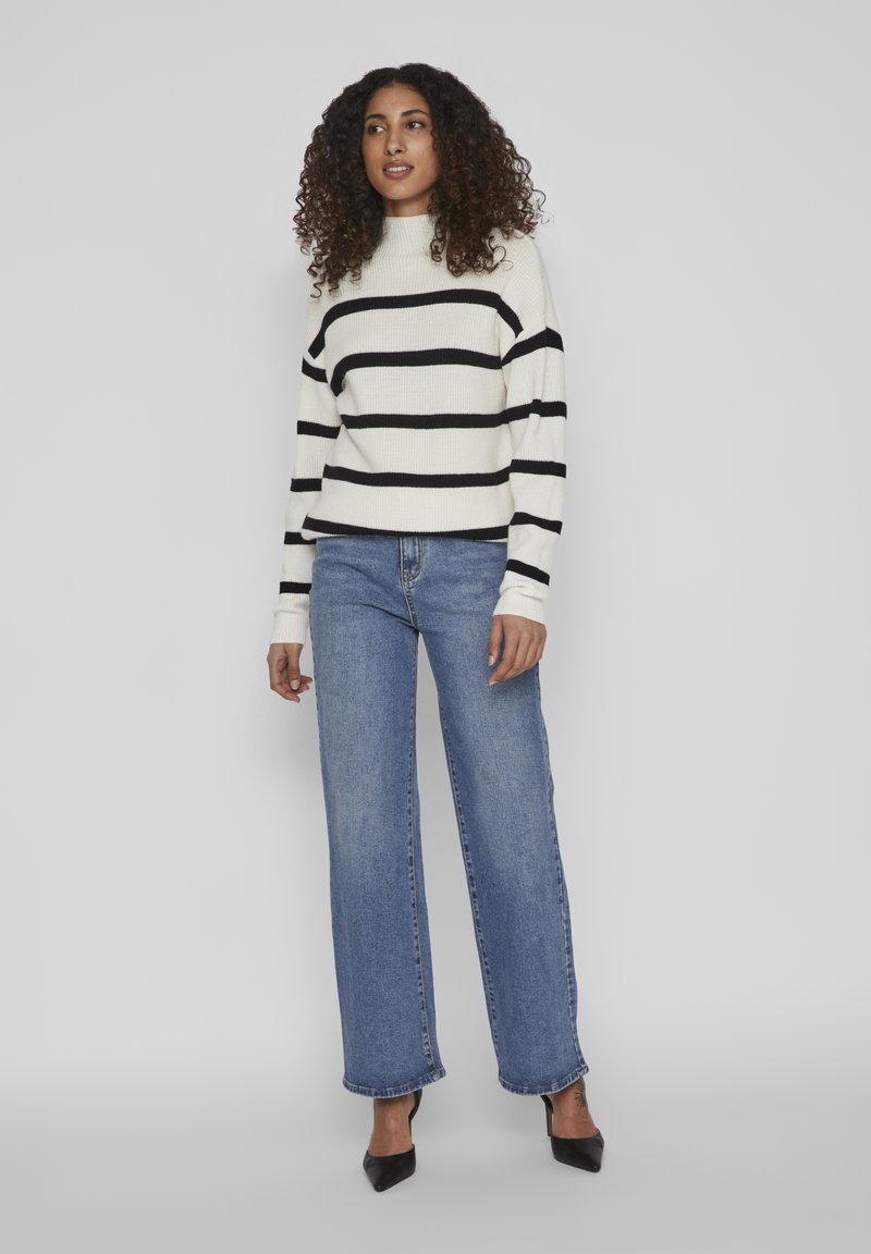 Pull rayé crème avec des lignes noires, col rond et texture côtelée ; associé à un jean bleu à jambes larges et des talons aiguilles noirs.