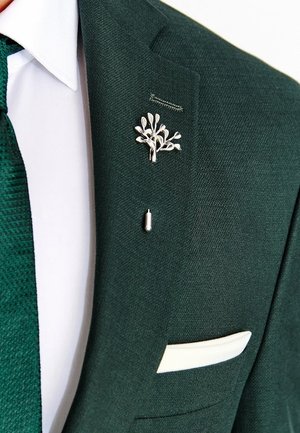 Ciemnozielona marynarka garniturowa z białą koszulą, teksturowanym zielonym krawatem, srebrną przypinką w kształcie drzewa na klapie oraz białą poszetką.