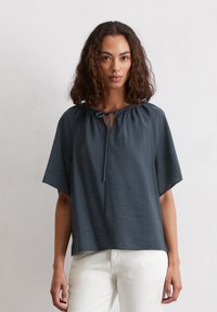 Maglietta grigia scura a maniche corte con collo rotondo e dettaglio con nodo frontale. Tessuto morbido e leggero con una vestibilità rilassata, abbinata a pantaloni color crema.