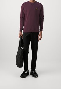 Sudadera granate con cuello redondo y logo amarillo, combinada con pantalones negros y bolso tote acolchado negro. Completan el conjunto unas zapatillas negras.