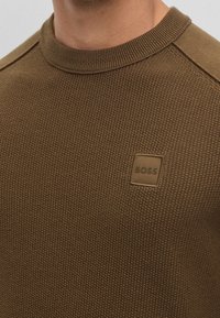 Brun stickad tröja med en texturerad yta, rund halsringning och en rektangulär logotyp på bröstet. Subtila axelaccenter.