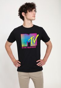 Black cotton t-shirt featuring a colorful MTV logo with a gradient design and the text "MUSIC TELEVISION" in pink. Model stands with hands on hips.