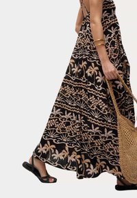 Abito maxi nero con motivi floreali e a onde beige, realizzato in tessuto leggero. Borsa in paglia intrecciata e sandali neri completano il look.