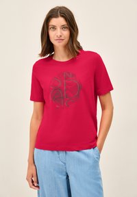 T-shirt en coton rouge à manches courtes, avec un motif graphique circulaire noir sur le devant. Texture douce et coupe ajustée.