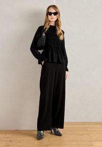 Černý peplum top s texturou a dlouhými rukávy, spárovaný s volnými černými kalhotami. Šedé lodičky na vysokém podpatku s bílými kruhovými akcenty a slunečními brýlemi.
