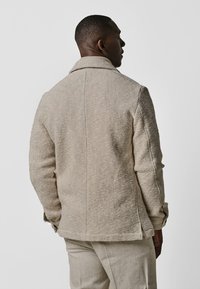 Beige strukturierte Jacke mit Kragen, sichtbaren Nähten und Seitentaschen. Getragen mit hellen Hosen, von hinten dargestellt.