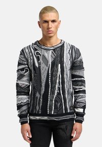 Pull en tricot noir et gris avec des motifs et textures complexes, doté de manches longues et d'un col rond. Les accents incluent une étiquette sur l'ourlet.