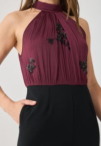 Top sans manches bordeaux avec un col montant, en tissu plissé, et des embellissements floraux noirs. Assortie à un bas noir uni.