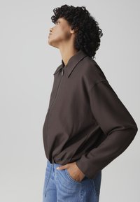 Persona con capelli ricci che indossa una giacca marrone scuro con zip e jeans azzurri, in piedi di profilo con la mano in tasca.