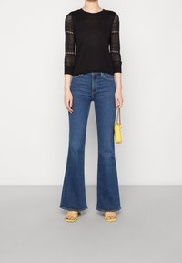 Czarna bluzka z długim rękawem ozdobiona koronką, w zestawie z dżinsami w kolorze niebieskim typu dzwon. Na nogach żółte sandały na paski; trzyma w ręku małą żółtą torebkę.