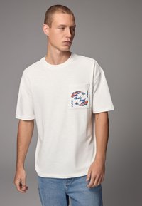 White short-sleeve t-shirt featuring a front pocket with colorful koi fish embroidery and "Honolulu" text, paired with light blue jeans.