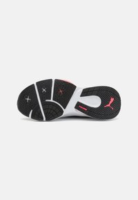 Suela de goma negra con patrones de agarre texturizados. La entresuela blanca presenta un acento rosa con el logo de 'PUMAGRIP'. Forma curva para un ajuste ergonómico.
