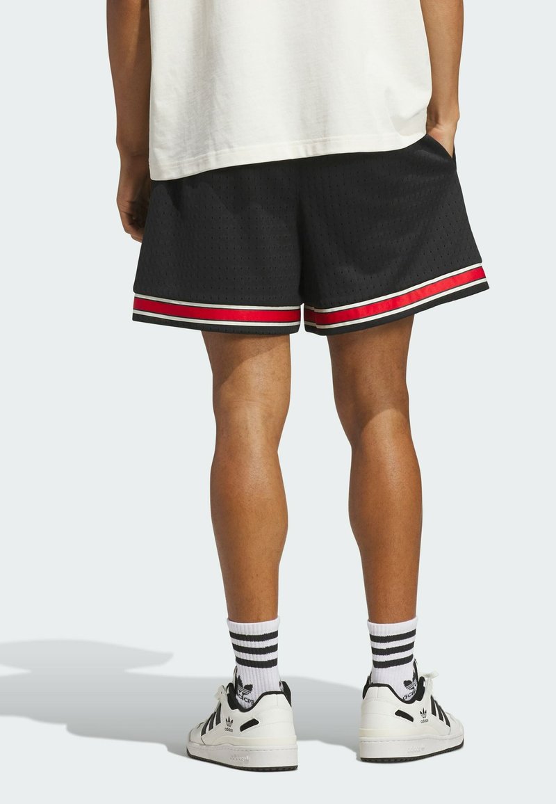 Person trägt schwarze Mesh-Shorts mit rotem und weißem Besatz, weiße Socken mit schwarzen Streifen und weiße Adidas-Sneakers, steht mit den Händen in den Taschen.