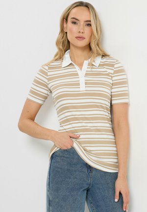 Frau trägt ein beige-weiß gestreiftes Poloshirt mit kurzen Ärmeln, blaue Jeans, eine Hand in der Tasche, steht vor weißem Hintergrund.