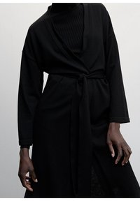 Sort langærmet slå-om-kjole med bundet talje, med dyb V-udskæring og glat tekstur, lagdelt over en ribbet sort top.