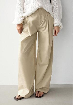 Person trägt beige weit geschnittene Hose, weißen gestrickten Pullover, silbernes Armband und braune Flip-Flops, steht drinnen vor einer schlichten Wand.