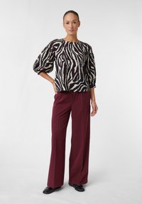 Schwarz-beige gestreifte Bluse im Zebra-Look mit Puffärmeln, kombiniert mit hoch taillierten burgunderfarbenen Hosen. Glatter Stoff und taillierte Passform.