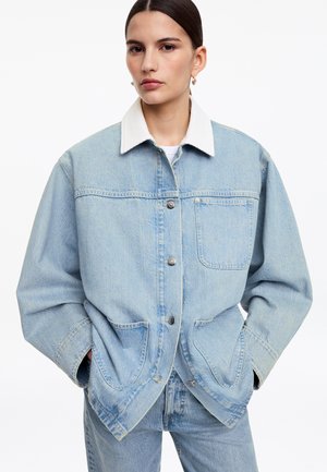 Femme aux cheveux raides et foncés porte une veste en denim bleu clair oversize avec col blanc, mains dans les poches, assortie à un jean en denim.