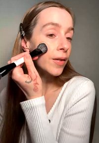 Femme aux longs cheveux vêtue de blanc, appliquant du maquillage avec un pinceau sur la joue, avec une pince tenant les cheveux en arrière et un tatouage de rose sur la main.