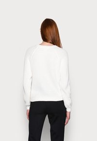 Witte gebreide trui met een ribbeltextuur, ronde hals, raglanmouwen en knoopdetails op de manchetten, gecombineerd met zwarte broeken.