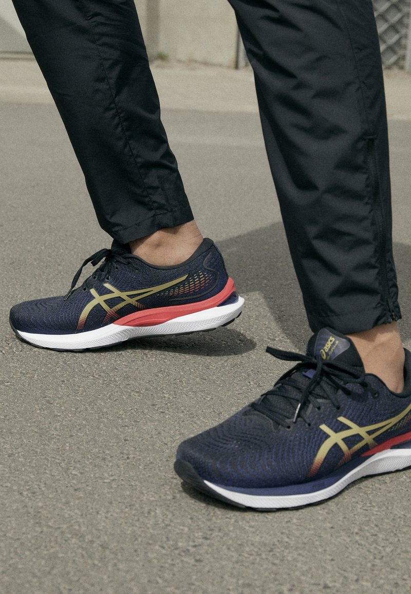 Henkilö kävelee jalkakäytävällä mustissa housuissa ja tummansinisissä juoksukengissä, joissa on kultaiset yksityiskohdat ja punaiset välipohjat.