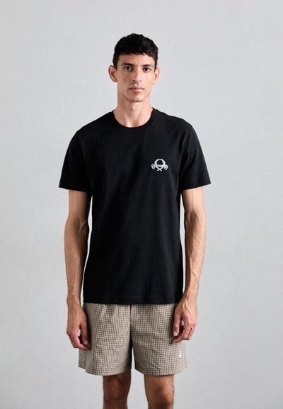 T-shirt en coton noir à manches courtes avec un petit motif blanc sur la poitrine. Porté avec un short à carreaux beige, mettant en valeur une tenue décontractée.