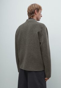 Giacca grigia in lana con una vestibilità rilassata, spalle ampie e orlo arrotondato. Presenta un design minimalista e una superficie liscia e testurizzata.