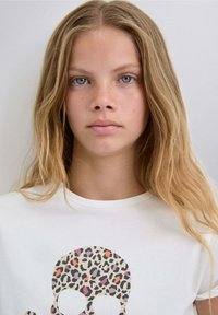 Camiseta blanca con un gráfico de una calavera en estampado de leopardo con manchas naranjas, rosas y negras. Cuello recto y textura suave.
