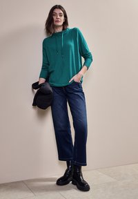 Türkisfarbener Kapuzenpullover mit strukturierten Streifen, kombiniert mit dunkelblauen weiten Jeans und schwarzen klobigen Stiefeln. Das Modell hält eine schwarze wattierte Jacke.
