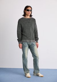 Gray knit sweater with ribbed texture, distressed hem, light blue jeans, cream sneakers with brown stripes, and black sunglasses.