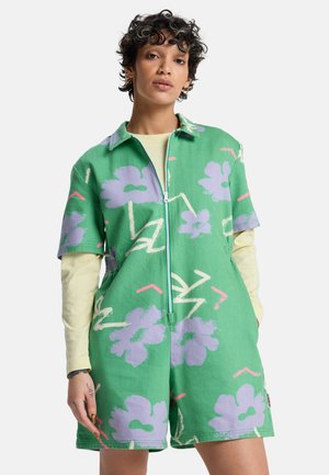 Person trägt grünen Kurzarm-Overall mit lila floralem und abstraktem Muster, Frontreißverschluss, über einem beigen Langarmshirt, posiert mit Händen in den Taschen.