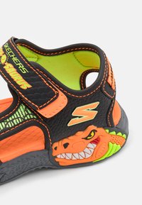 Svart sandaler med neongrønne detaljer, teksturert materiale og et dinosaurmotiv. Har justerbar stropp og en grønn såle.