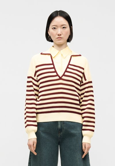 Femme aux cheveux noirs raides portant un pull à col en V à rayures crème et bordeaux par-dessus une chemise à col jaune pâle et un jean bleu.