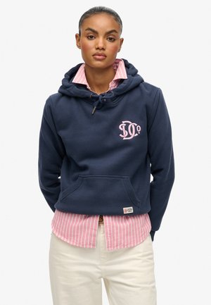 Dunkelblauer Kapuzenpullover mit einem pinkfarbenen Logo, Kordelzug am Hals und Tasche. Trägt sich über einem hellrosa-weiß gestreiften Hemd mit Kragen.