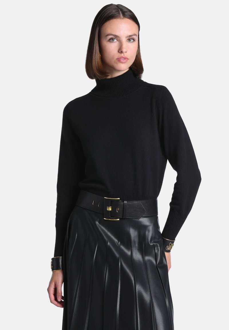 Femme aux cheveux bruns raides portant un pull à col roulé noir, une large ceinture noire avec une boucle dorée, et une jupe en cuir plissée noire.