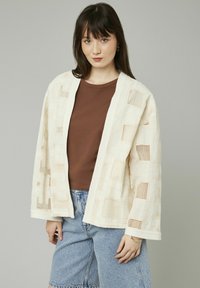 Gilet en crème avec un design tissé texturé présentant des motifs carrés ; porté sur un haut marron et associé à un short en denim bleu clair.